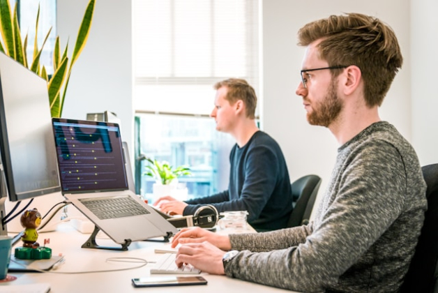 Deux juenes hommes travailent côte à côte dans un openspace à leur bureau, devant leurs ordinateurs protables